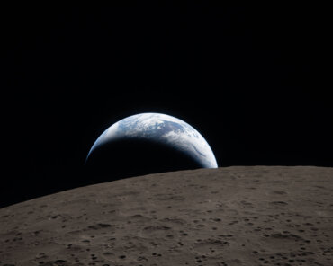 Earthset captured through the Orion spacecraft window at 6:41 p.m. EDT, April 6, 2026, during the Artemis II crew’s flyby of the Moon. A muted blue Earth with bright white clouds sets behind the cratered lunar surface.
The dark portion of Earth is experiencing nighttime. On Earth’s day side, swirling clouds are visible over the Australia and Oceania region.  
In the foreground, Ohm crater has terraced edges and a flat floor interrupted by central peaks. Central peaks form in complex craters when the lunar surface, liquefied on impact, splashes upwards during the crater’s formation. Image: NASA